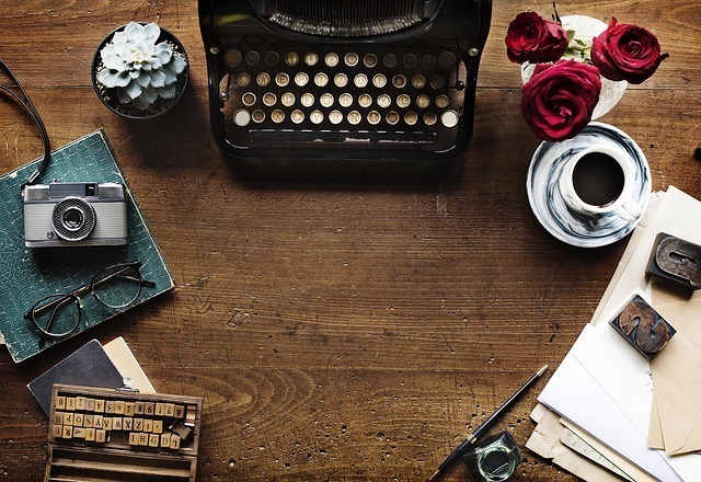 A desk with books, papers, a camera, two plants, coffee, stamps, and a typewriter.