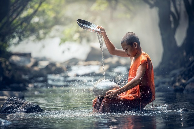 A young monk pours water on a metal drum as he sits in a river.